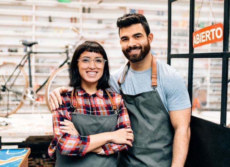 Couple in a workshop taking photos to be used on social media. The women has short hair and is wearing a checked shirt and apron which is grey, the man wears the same apron and a blue t-shirt and has brown hair