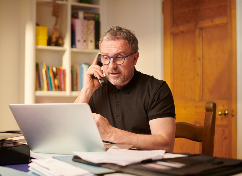 The business owner, male, 50, is on the phone in front of the laptop, relaxed and happy; he is wearing glasses.