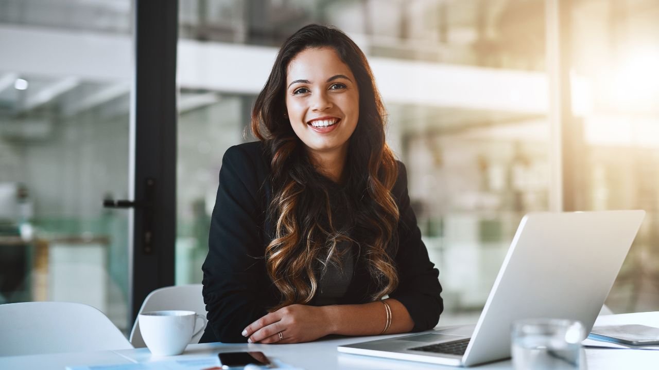 A professional woman sitting confidently at a desk with a laptop, smiling directly at the camera in a bright, modern office. The image conveys confidence, professionalism and readiness for the next opportunity.