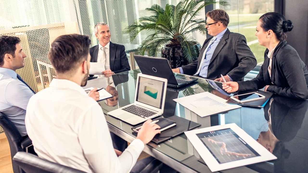 A group of professionals gathered around a conference table in a modern office, engaged in a business meeting. Several laptops, tablets, notebooks and printed reports are visible on the table, with the participants listening and contributing to the discussion. The image conveys collaboration, communication, decision making and the importance of being able to explain your experience and ideas in a professional setting.